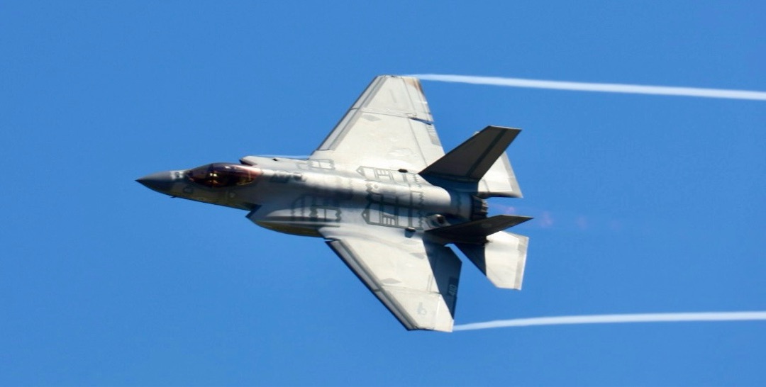An F-35 fighter jet banking against blue sky with vapor trails